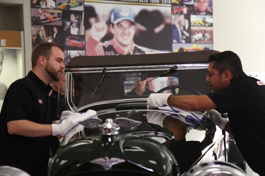 two men in black shirts wiping a dark green Duesenberg automobile with cloths.